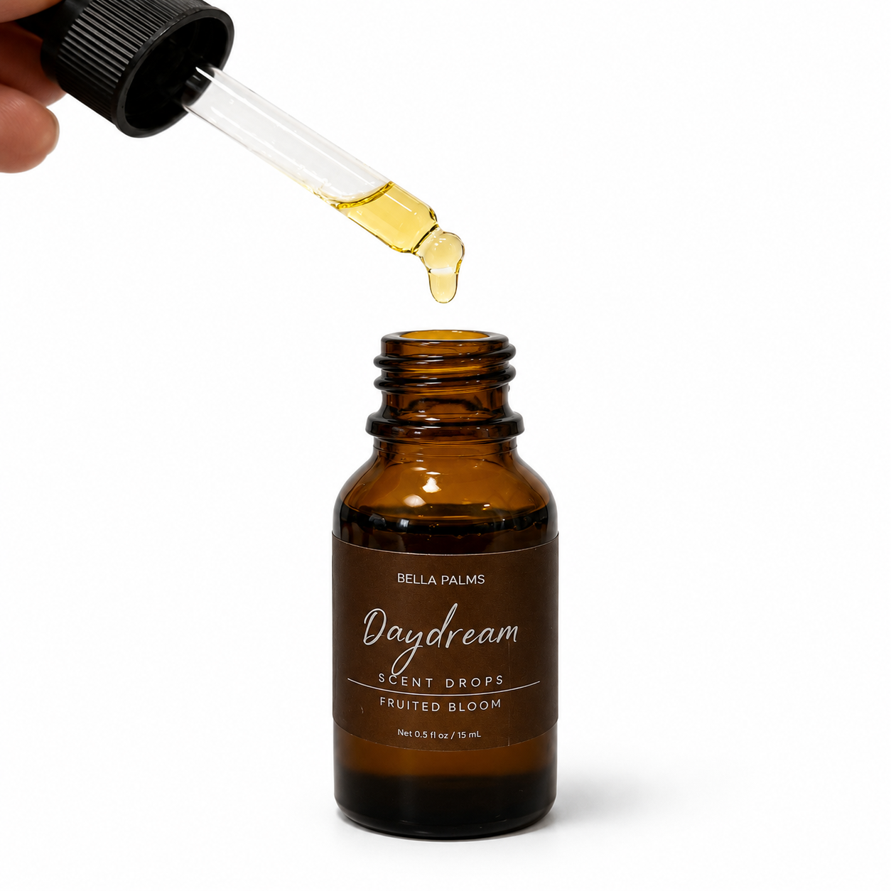 A hand holds a glass dropper above an amber glass bottle labeled Bella Palms Daydream Scent Drops, with a drop of golden-yellow liquid dripping into the bottle against a white background.